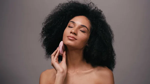 Brunette afro-américaine aux épaules nues utilisant un dispositif de nettoyage en silicone sur le visage isolé sur gris — Photo de stock