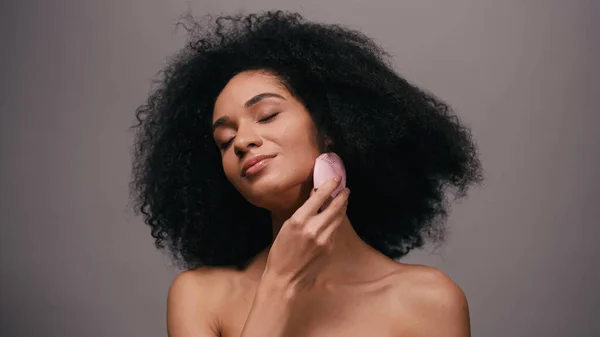 Femme afro-américaine heureuse avec les épaules nues en utilisant un dispositif de nettoyage en silicone sur le visage isolé sur gris — Photo de stock