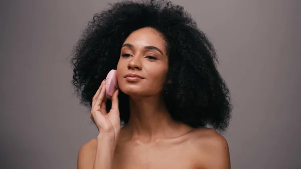 Молода афроамериканка з голими плечима, використовуючи пристрій для очищення обличчя ізольована на сірому — стокове фото