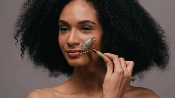 Pretty african american woman with bare shoulders using jade roller isolated on grey — Stock Photo