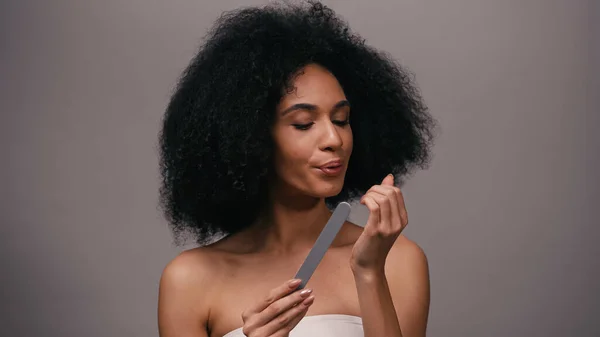 Giovane donna afro-americana che tiene la lima per unghie e soffia sulle unghie isolate sul grigio — Foto stock