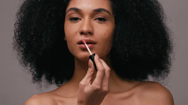 Morena mujer afroamericana aplicando brillo de labios y mirando a la cámara aislada en gris - foto de stock