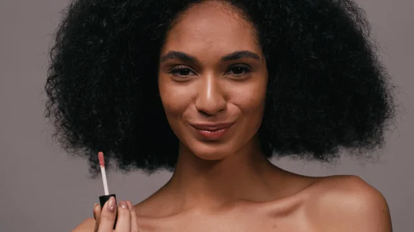Mujer afro-americana feliz con brillo de labios mirando a la cámara aislada en gris - foto de stock