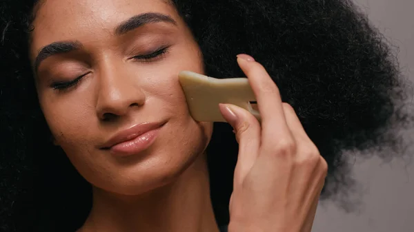 Curly african american woman using gua sha stone isolated on grey — Stock Photo
