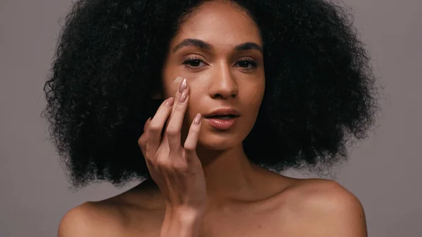 Mujer americana africana rizada aplicando la base de la cara con los dedos aislados en gris - foto de stock