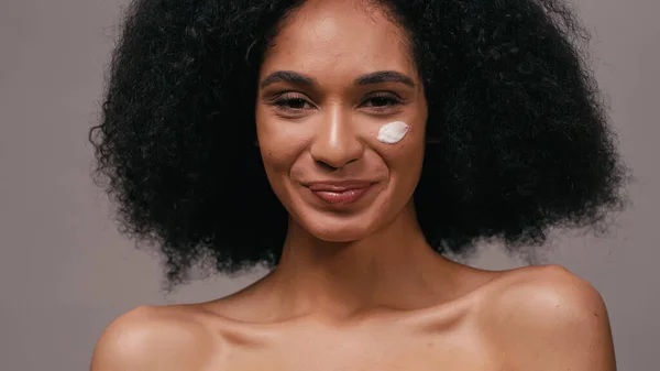 Alegre mujer afroamericana con crema cosmética en la cara aislada en gris - foto de stock