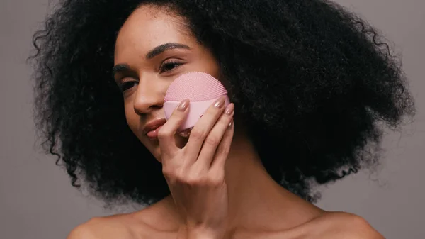 Encaracolado afro-americano mulher usando gadget de limpeza no rosto isolado em cinza — Fotografia de Stock