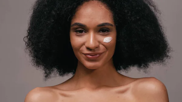 Femme afro-américaine souriante avec crème cosmétique sur le visage isolé sur gris — Photo de stock
