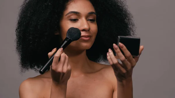 Mujer afroamericana aplicando polvo facial con cepillo cosmético aislado en gris - foto de stock