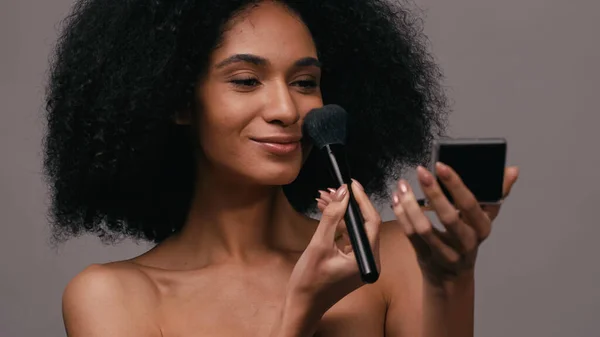 Mujer afroamericana complacida aplicando polvo facial con cepillo cosmético aislado en gris - foto de stock