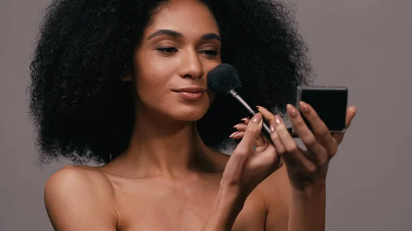 Mujer joven afroamericana aplicando polvo facial con cepillo cosmético aislado en gris - foto de stock