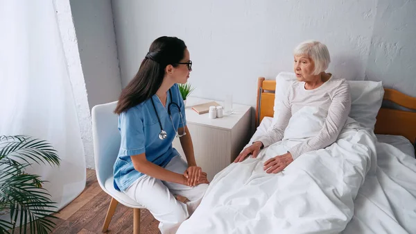 Enfermera joven en anteojos y mujer anciana hablando en la clínica - foto de stock