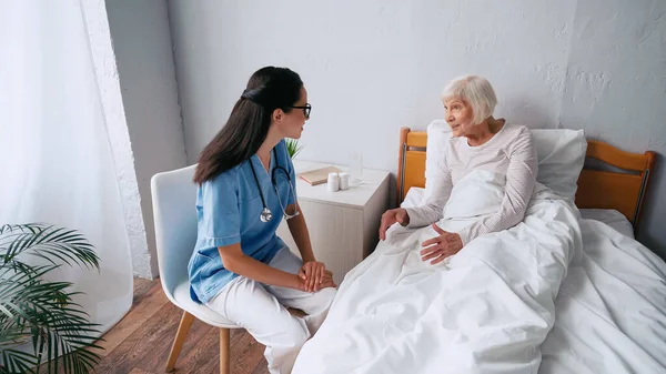 Медсестра в очках и улыбающаяся пожилая женщина разговаривающая в клинике — стоковое фото