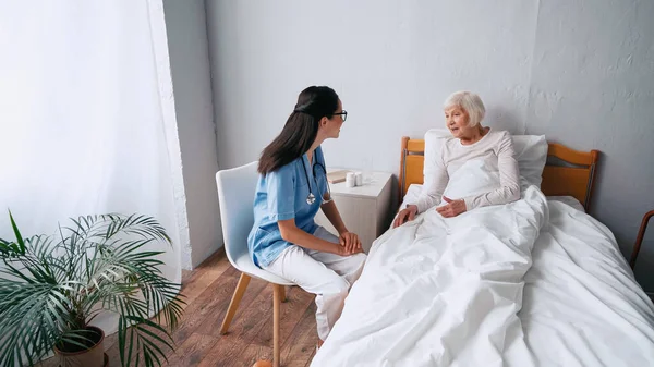 Enfermera alegre en anteojos y anciana hablando en clínica - foto de stock