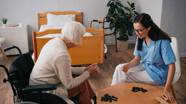 Senior femme en fauteuil roulant jouer dominos avec heureux infirmière — Photo de stock