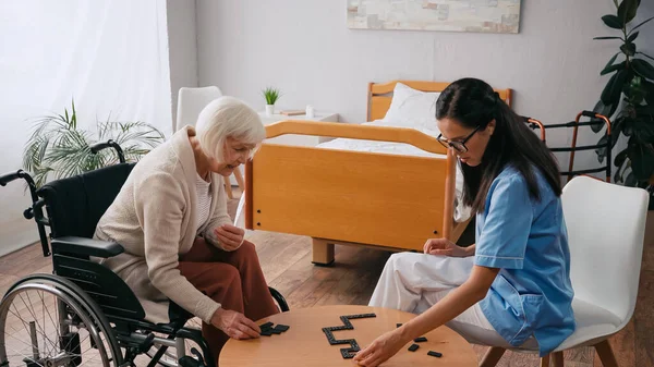 Seniorin im Rollstuhl spielt Dominosteine mit junger Krankenschwester — Stockfoto