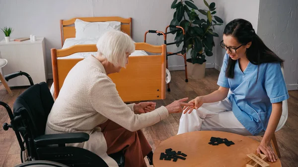 Senior femme en fauteuil roulant jouer dominos avec brunette infirmière — Photo de stock