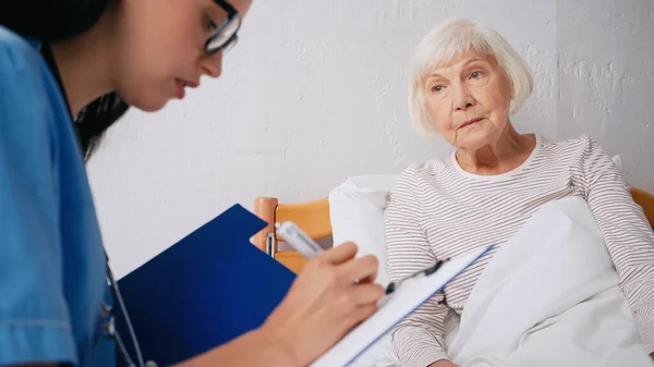 Enfermera borrosa en anteojos escribir prescripción cerca de la mujer anciana - foto de stock