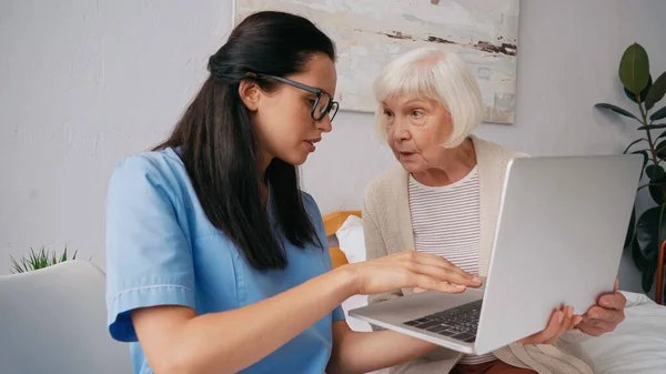 Enfermeira geriátrica morena mostrando mulher idosa como usar laptop — Fotografia de Stock