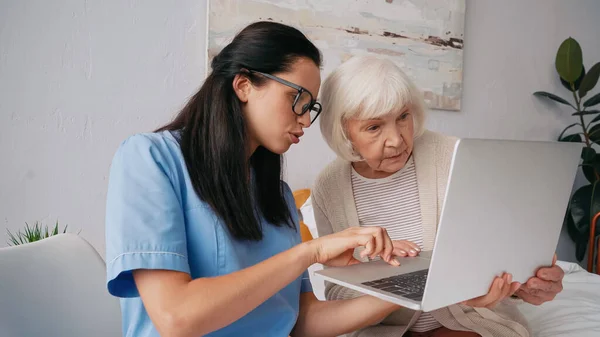 Enfermera morena mostrando a la mujer anciana cómo utilizar el ordenador portátil - foto de stock