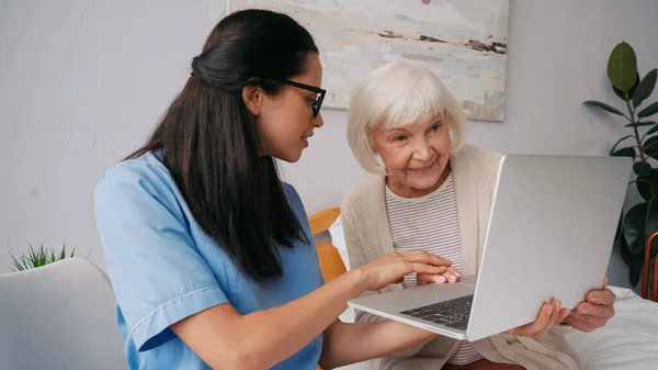 Bruna infermiera geriatrica mostrando felice donna anziana come utilizzare il computer portatile — Foto stock