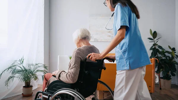 Enfermera geriátrica en anteojos moviendo a una mujer anciana en silla de ruedas - foto de stock