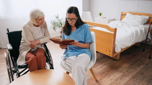 Joven geriatra leyendo libro a una mujer anciana en silla de ruedas - foto de stock