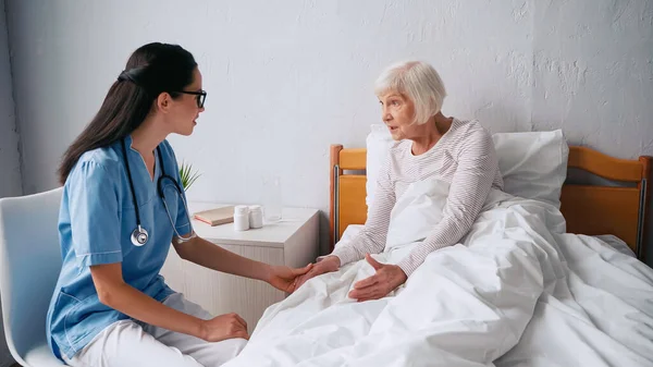 Enfermera geriátrica en anteojos hablando con mujer anciana en la cama - foto de stock
