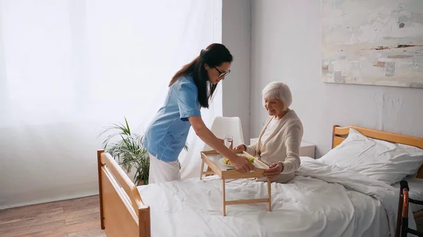 Brünette Krankenschwester trägt Tablett mit Frühstück zu lächelndem älteren Patienten — Stockfoto