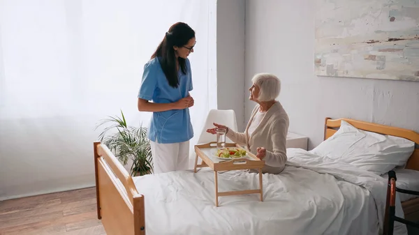 Enfermeira morena conversando com paciente idoso alegre perto bandeja de café da manhã na cama — Fotografia de Stock