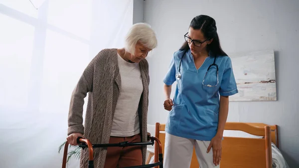 Senior woman stepping with walkers near brunette nurse in eyeglasses — Stock Photo