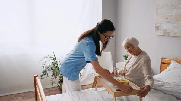 Cheerful nurse bringing tray with breakfast to aged patient — Stock Photo