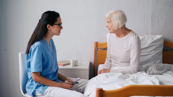 Сестра брюнетка разговаривает с счастливым пожилым пациентом и держит стетоскоп — стоковое фото