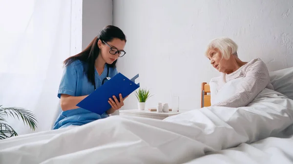 Брюнетка медсестра в очках выписывает рецепт в буфер обмена рядом с пожилой женщиной — стоковое фото