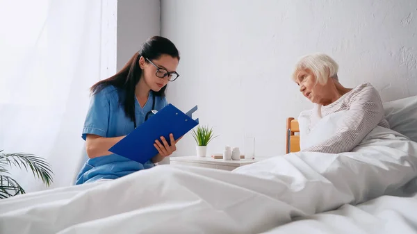 Enfermera morena en gafas graduadas escribir prescripción en portapapeles cerca de la mujer anciana - foto de stock