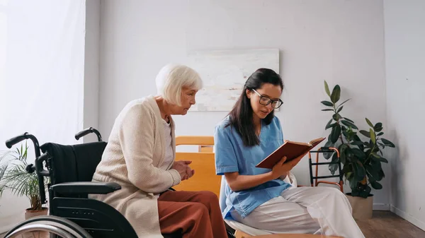 Brunette geriatrician nurse reading book to aged woman in wheelchair — Stock Photo