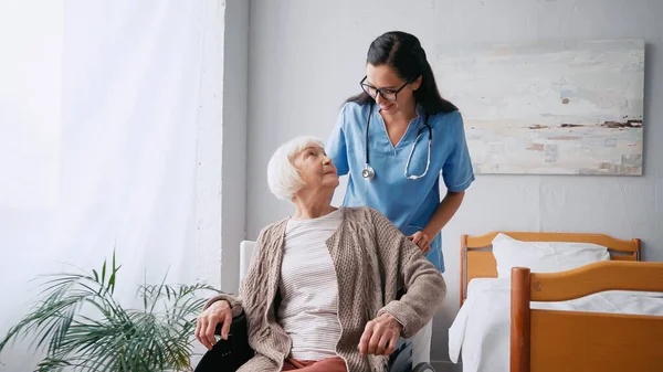 Enfermera feliz en gafas moviendo a mujer anciana en silla de ruedas - foto de stock