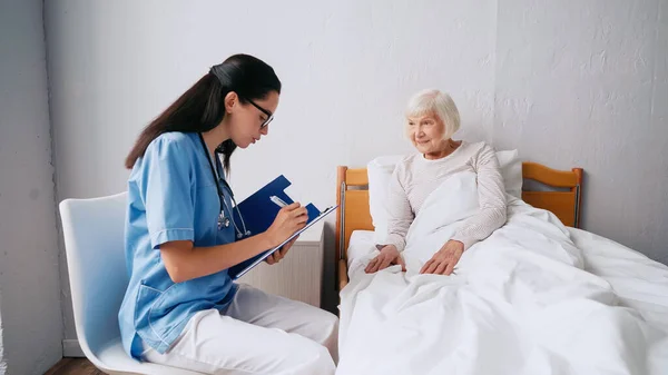 Infirmière brune dans les lunettes écrivant prescription dans le presse-papiers près de femme âgée au lit — Photo de stock