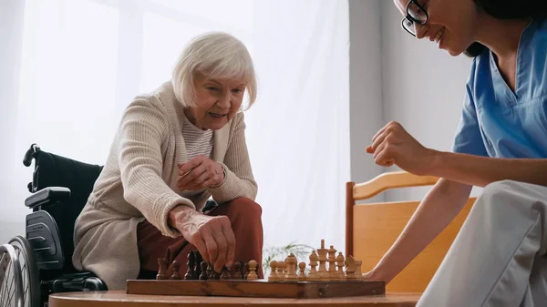 Літня жінка і щаслива медсестра грають в шахи в будинку престарілих — стокове фото