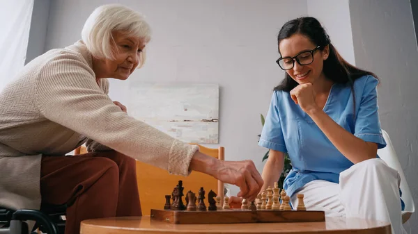 Anciana y enfermera alegre jugando ajedrez en el hogar de ancianos - foto de stock