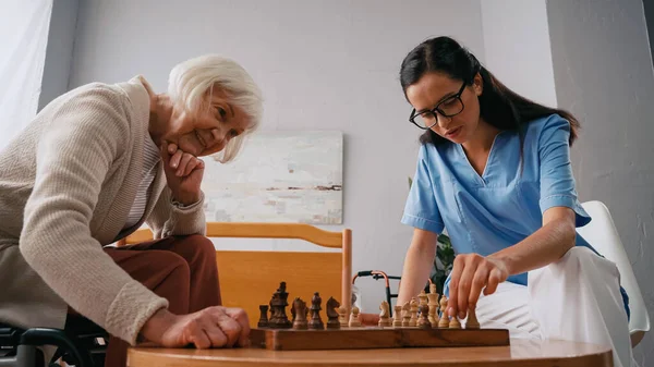 Feliz anciana y enfermera jugando al ajedrez en el hogar de ancianos - foto de stock