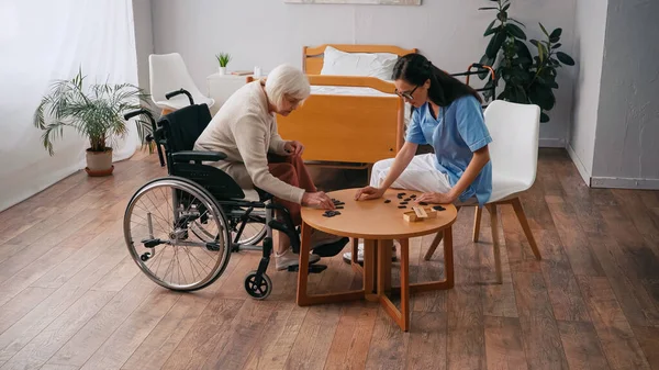 Anziana donna in sedia a rotelle giocare domino con bruna infermiera in occhiali — Foto stock