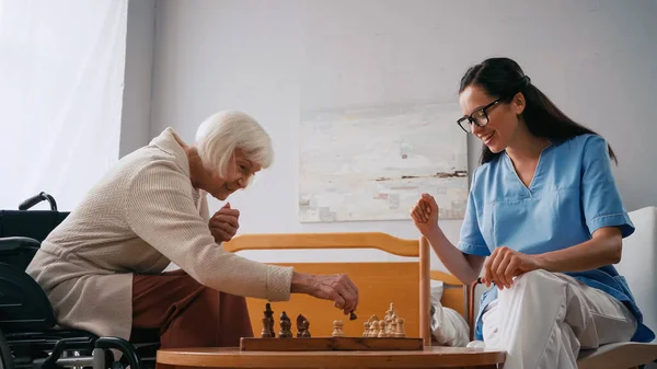 Happy elderly woman and smiling nurse playing chess in nursing home — Stock Photo