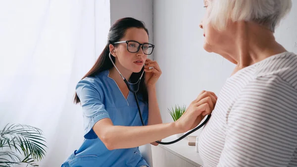 Брюнетка медсестра в очках осматривает пожилую женщину стетоскопом — стоковое фото