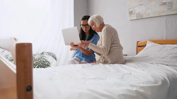 Брюнетка пожилая медсестра показывает пожилой женщине, как использовать ноутбук в доме престарелых — стоковое фото