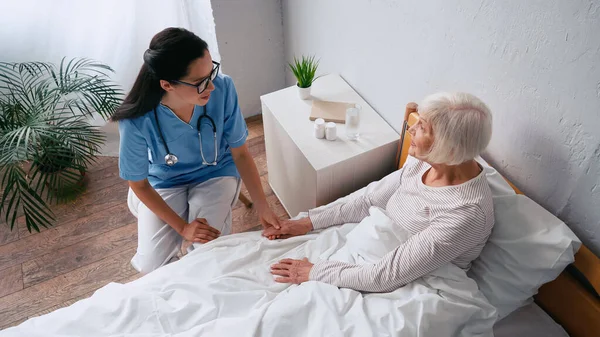 Vista de ángulo alto de enfermera morena en anteojos y mujer anciana feliz hablando en la clínica - foto de stock
