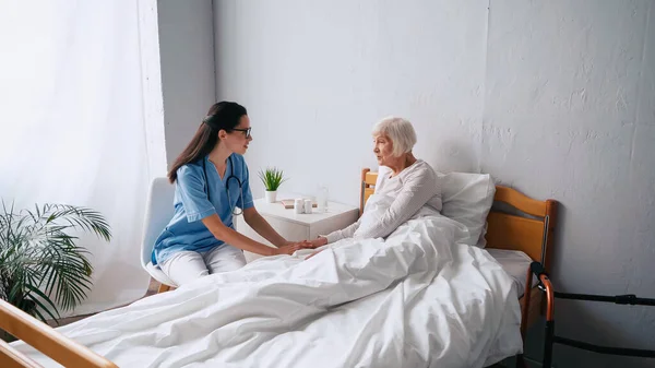 Brünette Krankenschwester mit Brille und Seniorin mit grauen Haaren reden in Klinik — Stockfoto