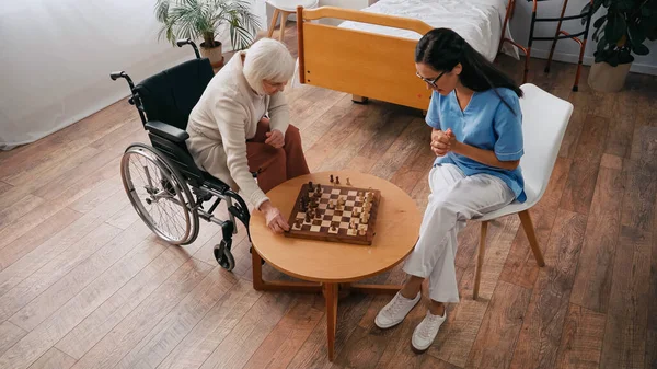 Visão de alto ângulo da mulher idosa e enfermeira jogando xadrez em casa de repouso — Fotografia de Stock