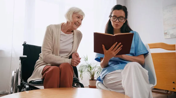 Brünette Altenpflegerin liest Buch für glückliche alte Frau im Rollstuhl — Stockfoto