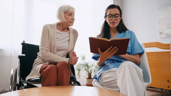 Brunette geriatrician nurse reading book to senior woman in wheelchair — Stock Photo